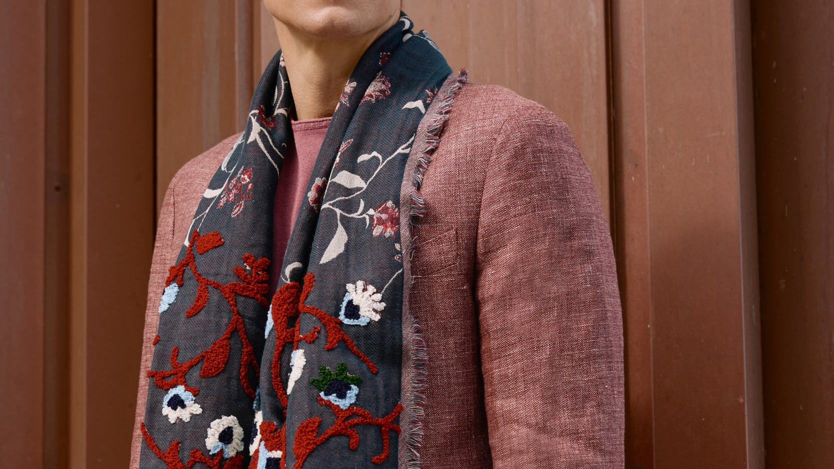 Man wearing a patterned scarf and pink jacket against a brown background